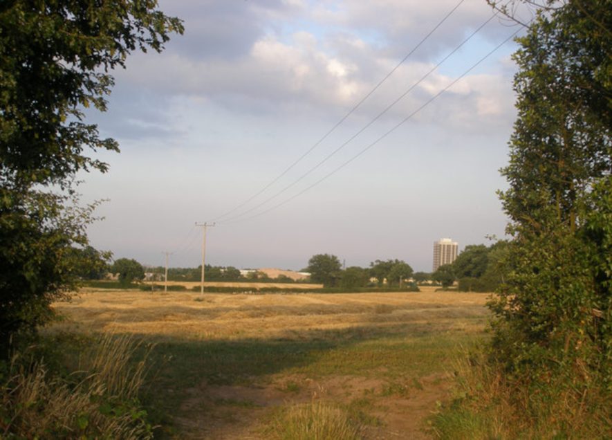 A dry flat field between hedgerows and trees with a tower block in the distance | Photo © John Evans (cc-by-sa/2.0). Originally uploaded to geograph.org.uk