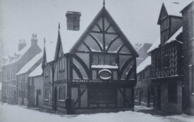 Black and white photograph of William Clarke's shop, with the words W. Clarke Baker visible on the side. It is winter, there is snow on the ground a roofs of the buildings. The building also known as Thomas Oken's House. It is a black and white timber framed building with windows made from small diamond shaped pieces of glass. | Warwickshire County Record Office reference PH1035/C1105