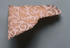Piece of wallpaper from Mary of Teck sent to Lady Eva Greville in a letter. The small piece of wallpaper has light pink background that shimmers. There is a pattern of scrolling leaves in a darker, matt, pink. | Warwickshire County Record Office reference CR1886/469/2/117