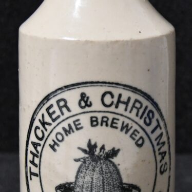 Stoneware ginger beer bottle sold by Thacker and Christmas. The label on the front of the bottle says this was home brewed ginger bear and has a drawing of a Christmas pudding in the centre. There is a sprig of holly on the top of the Christmas pudding. | Warwickshire Museum object reference H14903_1