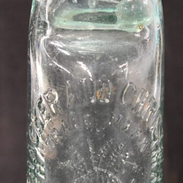 Glass Thacker and Christmas pop bottle. The bottle is made from a thick glass. It is hard to see in the image but the words Thacker and Christmas Warwick a raised relief on the bottle. An image of a Christmas pudding is also in raised relief on the side of the bottle. The marble stopper is missing from this bottle. | Warwickshire Museum object reference H19040