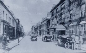 Photograph of Warwick High Street, showing the Warwick Arms Hotel. The photograph is in black and white. The images looks down the High Street towards the Westgate. The Warwick Arms Hotel is on the right hand side of the image. Who cars are parked outside the hotel. They appear to be early cars from the 1920s. This is probably when the photograph was taken. There are people in the image, walking on the pavement either side of the road, going about their daily business. The hotel has a canopy over the entrance that covers the pavement. One of the cars is parked in front of the canopy. Warwickshire museum document reference PH1035 C6033 B3209. | Warwickshire County Record Office reference PH1035/C6033/B3209.